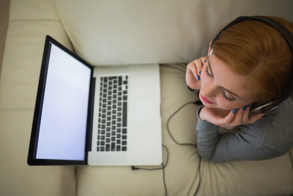 Redhead lying on the couch with her laptop wearing headphones at home in the sitting room